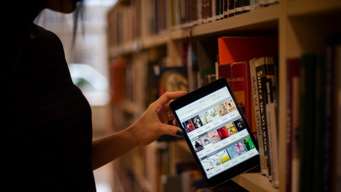 Sieviete bibliotēkas grāmatu plauktā ievieto planšeti ar 3td.lv e-grāmatu bibliotēkas skatu