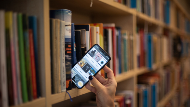 Sievietes roka izņem viedtālruni ar atvērtu 3td.lv skatu no bibliotēkas grāmatplaukta