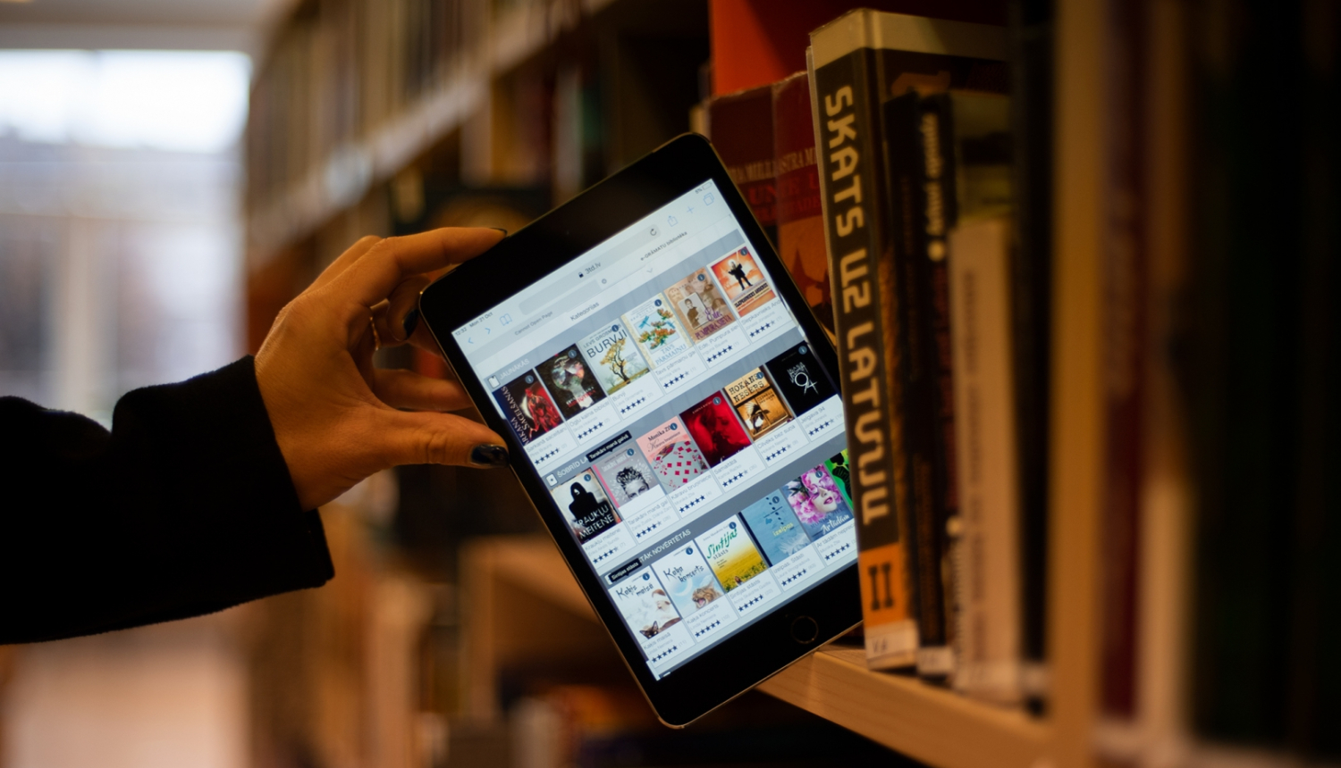 Planšetdators ar 3td bibliotēkas skatu publiskās bibliotēkas plauktā starp papīra grāmatām