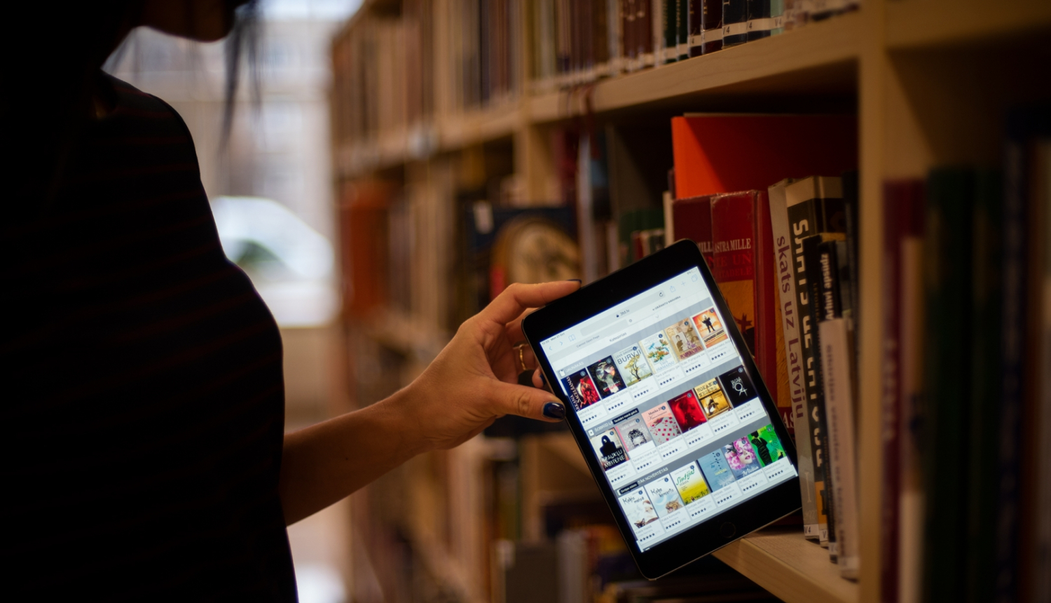 Sieviete bibliotēkas grāmatu plauktā ievieto planšeti ar 3td.lv e-grāmatu bibliotēkas skatu