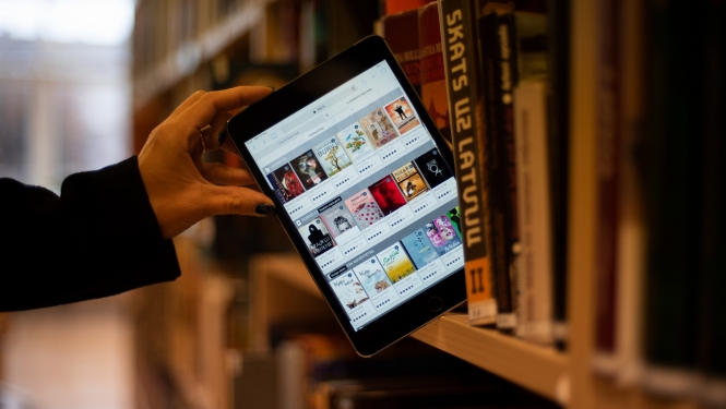 Planšetdators ar 3td bibliotēkas skatu publiskās bibliotēkas plauktā starp papīra grāmatām
