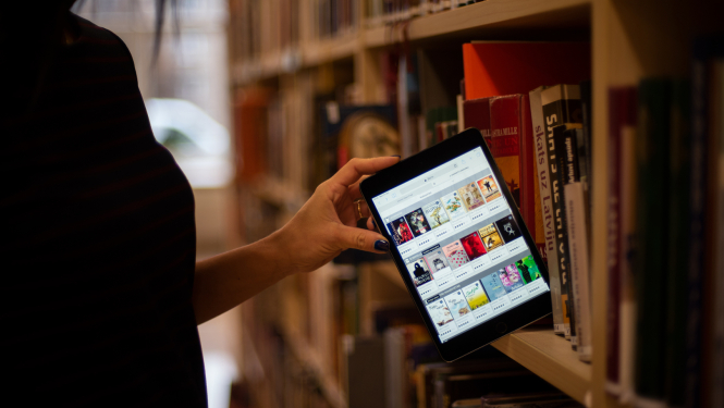 Sieviete ievieto bibliotēkas grāmatplauktā planšeti ar atvērtu 3td.lv skatu