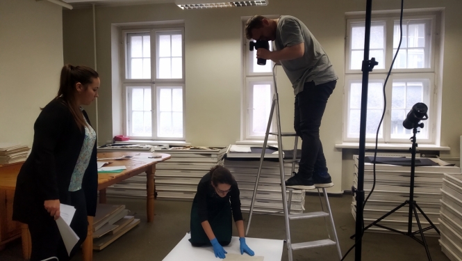 Fotogrāfs stāv uz trepēm un fotografē no augšas muzeja priekšmetu-dokumentu