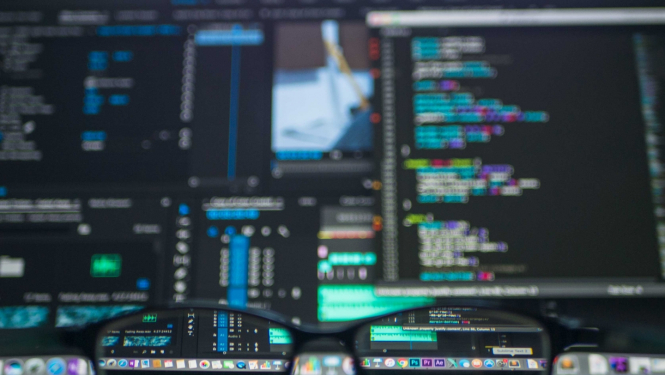 Vairāki datora ekrāni ar kodu sarakstiem un brilles priekšplānā uz galda