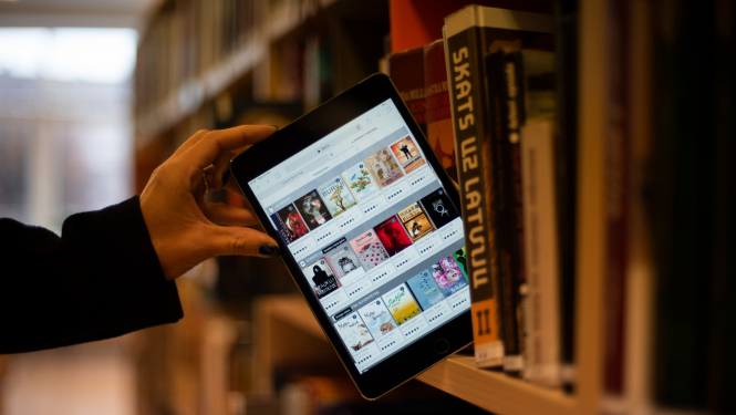 Planšetdators ar 3td bibliotēkas skatu publiskās bibliotēkas plauktā starp papīra grāmatām