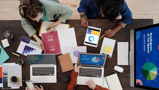 Skats no augšas uz cilvēkiem kas pie galda strādā pie laptopiem ar dažādām diagrammām