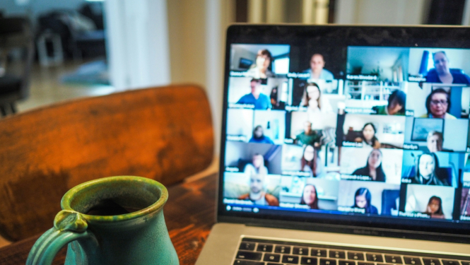 Atvērts portatīvais dators ar tiešsaistes sanāksmi ekrānā un māla krūzi blakus uz galda