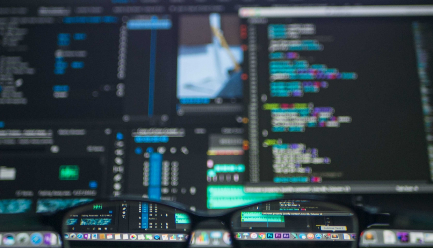 Vairāki datora ekrāni ar kodu sarakstiem un brilles priekšplānā uz galda