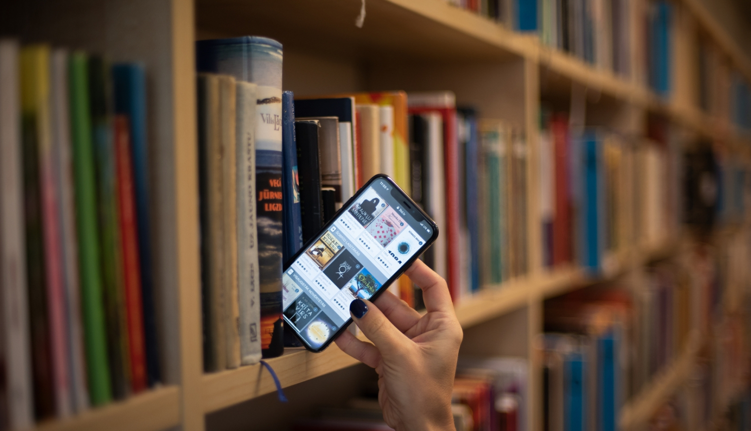 Sievietes roka izņem viedtālruni ar atvērtu 3td.lv skatu no bibliotēkas grāmatplaukta